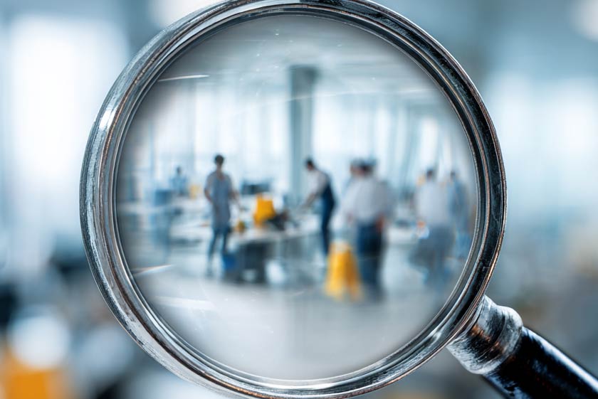 magnifying glass over a blurred image of a 3 cleaning people in a modern office space, illustrating lack of transparency